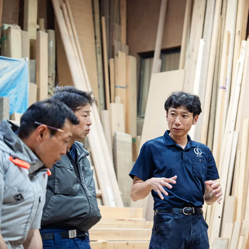 材木に囲まれた作業場で打ち合わせをする大工たち